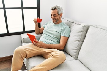 Wall Mural - Middle age grey-haired man using touchpad drinking coffee sitting on sofa at home