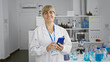 © Krakenimages.com - Blonde middle age woman scientist, all smiles as she efficiently uses her smartphone amidst work in the lab.