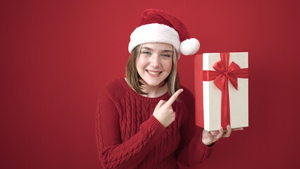 Wall Mural - Young blonde woman wearing christmas hat pointing with finger to gift over isolated red background