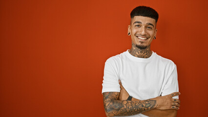 Confident latin young man, casually tattooed, poses with arms crossed, smiling at camera with joy, isolated over red wall background