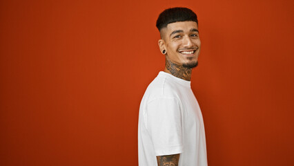 Cheerful young latin man smiling and looking confident, casually standing over isolated red background, exuding positive energy, happiness, and a cool lifestyle