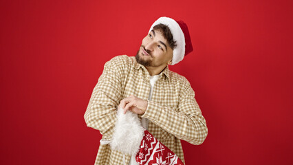 Canvas Print - Young arab man wearing christmas hat looking inside of sock over isolated red background