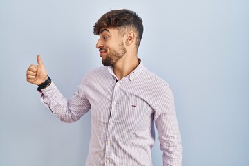 Wall Mural - Arab man with beard standing over blue background looking proud, smiling doing thumbs up gesture to the side