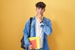 Hispanic teenager wearing student backpack and holding books with hand on chin thinking about question, pensive expression. smiling and thoughtful face. doubt concept.