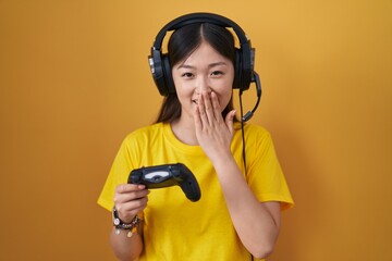 Wall Mural - Chinese young woman playing video game holding controller laughing and embarrassed giggle covering mouth with hands, gossip and scandal concept