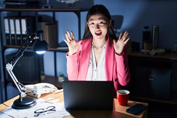 Canvas Print - Chinese young woman working at the office at night celebrating crazy and amazed for success with arms raised and open eyes screaming excited. winner concept