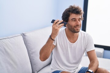 Poster - Young hispanic man listening voice message by smartphone sitting on sofa at home