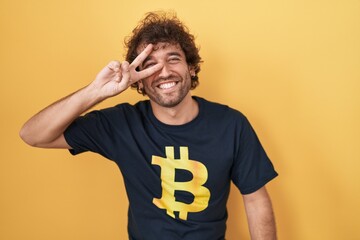 Sticker - Hispanic young man wearing bitcoin t shirt doing peace symbol with fingers over face, smiling cheerful showing victory