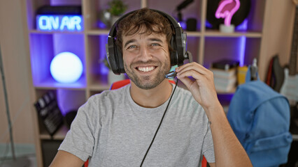 Happy young hispanic man streamer dons his futuristic gamepad and headphones, confidently streaming a cyber game, sitting at a table in his gaming room, smiling wide into the night.