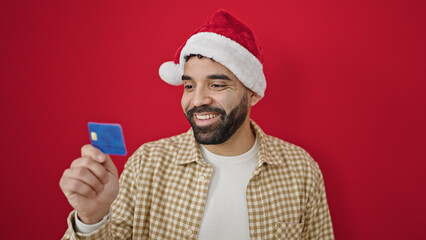 Sticker - Young hispanic man wearing christmas hat holding credit card over isolated red background