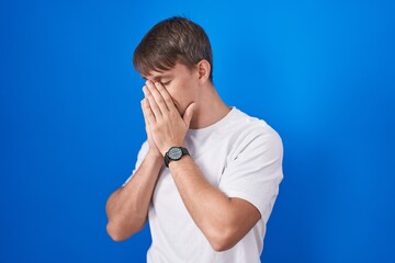 Wall Mural - Caucasian blond man standing over blue background with sad expression covering face with hands while crying. depression concept.