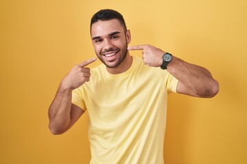 Wall Mural - Young hispanic man standing over yellow background smiling cheerful showing and pointing with fingers teeth and mouth. dental health concept.