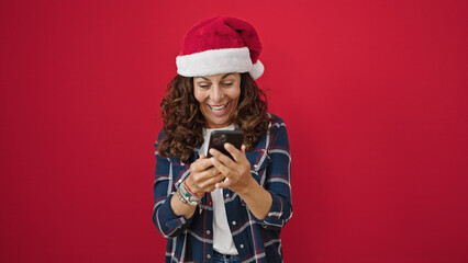 Canvas Print - Middle age hispanic woman wearing christmas hat using smartphone over isolated red background