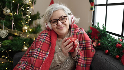 Wall Mural - Middle age woman with grey hair listening to music drinking coffee sitting by christmas tree at home