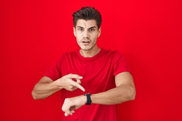 Poster - Young hispanic man standing over red background in hurry pointing to watch time, impatience, upset and angry for deadline delay