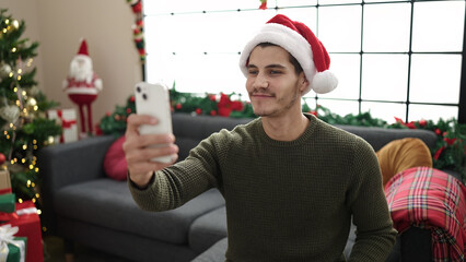 Wall Mural - Young hispanic man make selfie by camera sitting on sofa by christmas tree at home