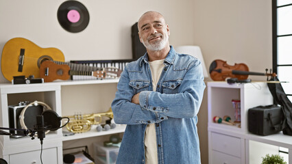 Wall Mural - Smiling middle-aged bald man with beard in a denim jacket standing in a music studio with instruments and recording equipment.