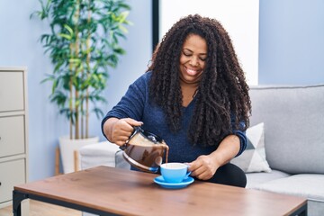 Canvas Print - African american woman pouring coffee on cup sitting on sofa at home