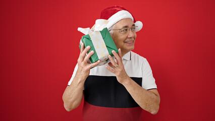 Wall Mural - Middle age man with grey hair smiling wearing christmas hat listening inside of gift over isolated red background