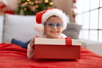 Sticker - Adorable hispanic toddler lying on sofa by christmas tree holding gift at home