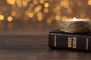 Candle burning and closed holy bible book on wooden table with bokeh background. Close-up. Copy space. Spiritual light shining in the darkness, Christian biblical concept.