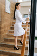 © dikushin - Vertical portrait of confident female doctor in medical gown standing posing on staircase at modern clinic. Pretty young woman professional doctor physician pediatrician standing in hospital.