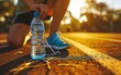Woman tying her shoes before a run on a racetrack in the park with bottle of water in focus on a bright background, with a blurred image of a sporty.