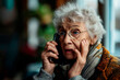 Worried elderly lady in eyeglasses talking on mobile phone, indoors with blurred background