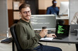 © Seventyfour - Medium long shot of smiling Caucasian male programmer in glasses looking at camera and holding takeaway coffee cup while sitting at desk in cybersecurity department