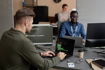 Poster - Male programmer writing software code using laptop while sitting at table in cybersecurity department, colleagues working in defocused background