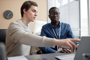 waist up shot of young caucasian male programmer pointing at laptop screen and talking to african am