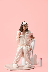 Poster - Young woman in pajamas with coffee using tablet computer on toilet bowl against pink background