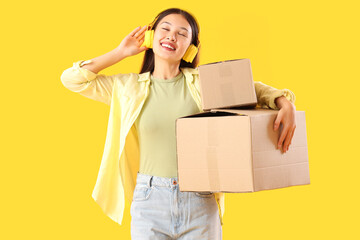 Sticker - Young Asian woman in headphones with parcels on yellow background