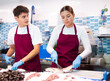 Proficient marketers in vinous aprons handling fresh seabass by scaler in fish shop