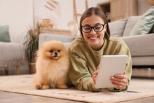 Young Woman With Cute Pomeranian Dog Using Tablet Computer On Floor At Home