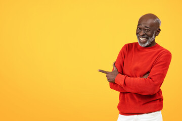 Wall Mural - Cheerful senior Black man with a white beard pointing to the side, wearing a bright red sweater