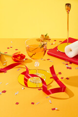 Wall Mural - An elegant glass of wine placed on a golden dish and decorated with a flower. A lot of confetti scattered on yellow surface with red ribbon and a tulip flower. Party celebration