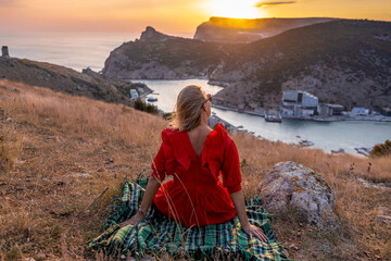 Wall Mural - Woman sunset sea mountains. Happy woman siting with her back on the sunset in nature summer posing with mountains on sunset, silhouette. Woman in the mountains red dress, summer landscape