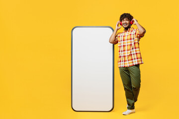 Wall Mural - Full body young happy Indian man he wears shirt casual clothes big huge blank screen mobile cell phone smartphone with mockup area listen music in headphones isolated on plain yellow color background.