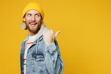 Wall Mural - Young smiling happy fun blond man he wears denim shirt hoody beanie hat casual clothes point thumb finger aside on area mockup isolated on plain yellow background studio portrait. Lifestyle concept.