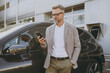 © ViDi Studio - Satisfied confident adult man customer male buyer client wearing white shirt classic grey suit standing near black electric car while charging talk speak on mobile cell phone outside Business concept