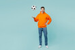 Full body young excited man fan wearing orange hoody casual clothes cheer up support football sport team hold in hand toss up soccer ball watch tv live stream isolated on plain blue color background.