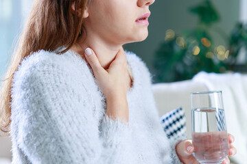 Wall Mural - Sick woman holding a glass of water touches her neck and feels sore throat due to flu