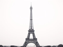 A Tall Metal Tower With A Large Arch With Eiffel Tower In The Background