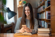 © Vitalii - Happy smiling university student girl using her mobile phone. Student working in university library, sitting at the desk with books and her phone