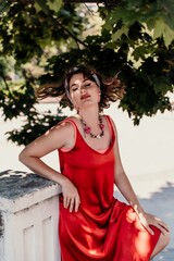 Poster - woman in a red silk dress and a bandage on her head smiles against the background of the leaves of a tree. She is leaning on the coop and looking into the camera. Vertical photo.