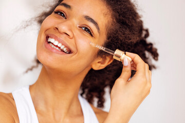 Cute smiling african young woman applies moisturizing serum from pipette to her cheek