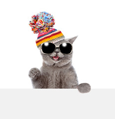Poster - Happy cat wearing sunglasses and knitted warm woolen hat with pompon looks above empty white board. isolated on white background