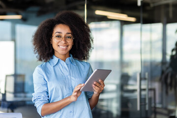 A young, beautiful businesswoman with curly hair working inside an office, holding a tablet.