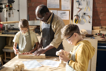 Portrait of senior craftsman teaching children carpentry in workshop lit by sunlight, copy space